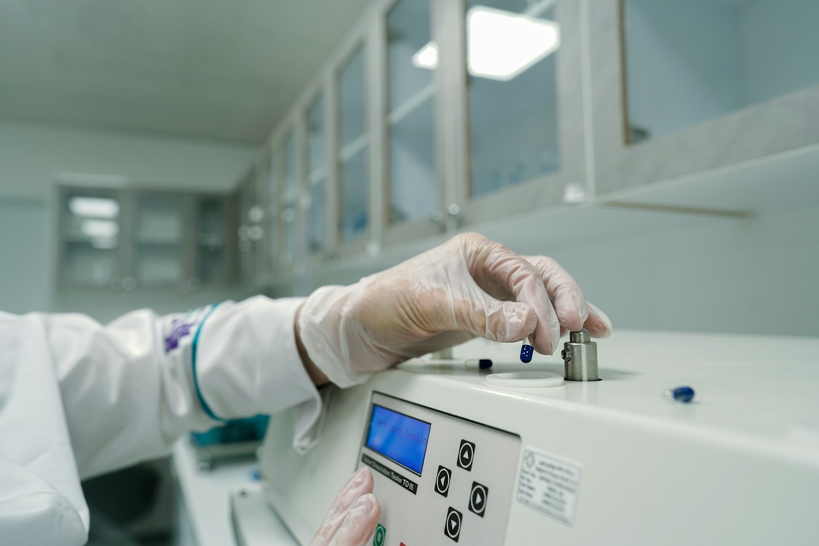 A gloved hand placing a blue capsule into a testing instrument with a small display and keypad in a pharmaceutical laboratory