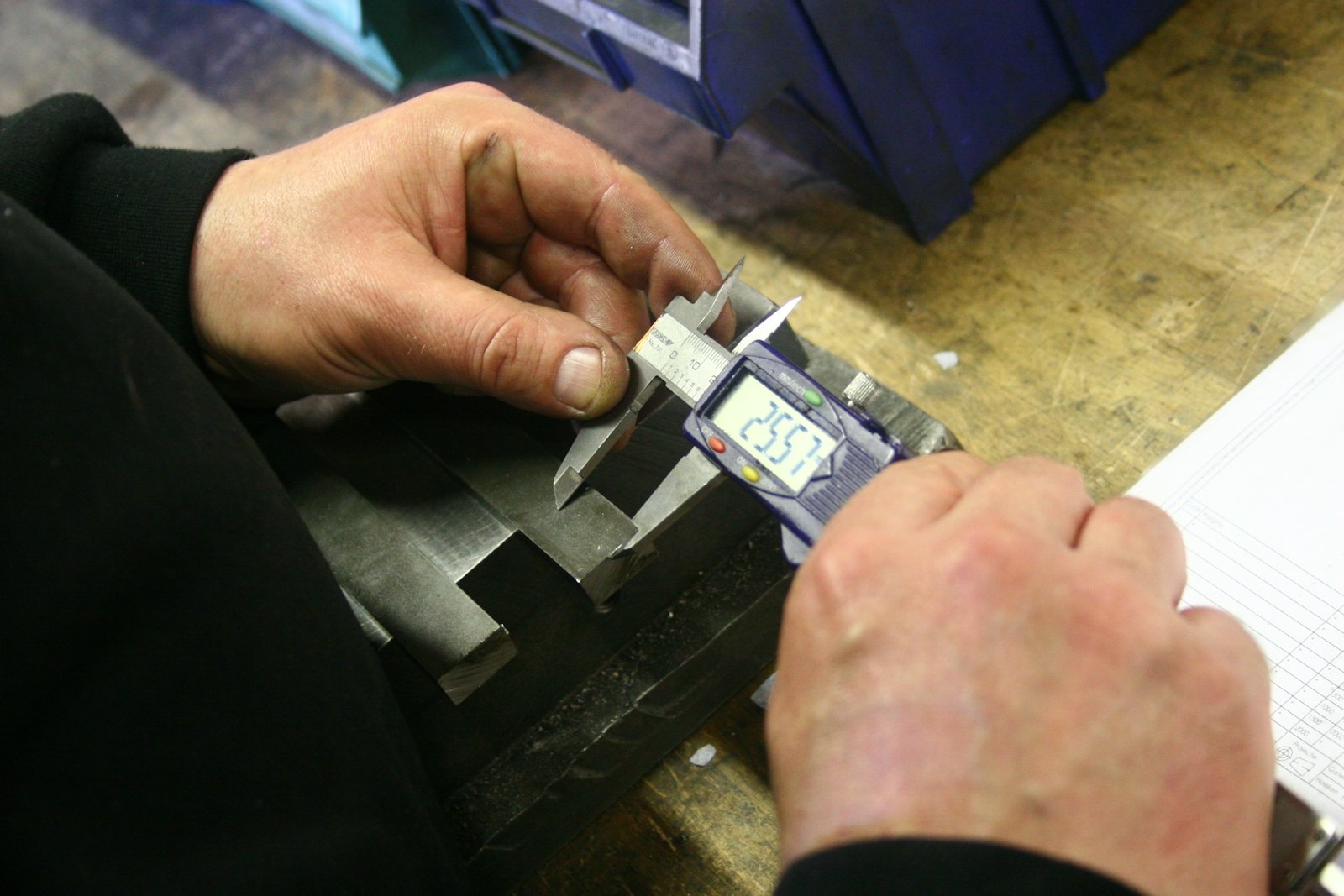 Close-up of two hands measuring a machined metal part with a digital caliper beside a printed inspection record sheet