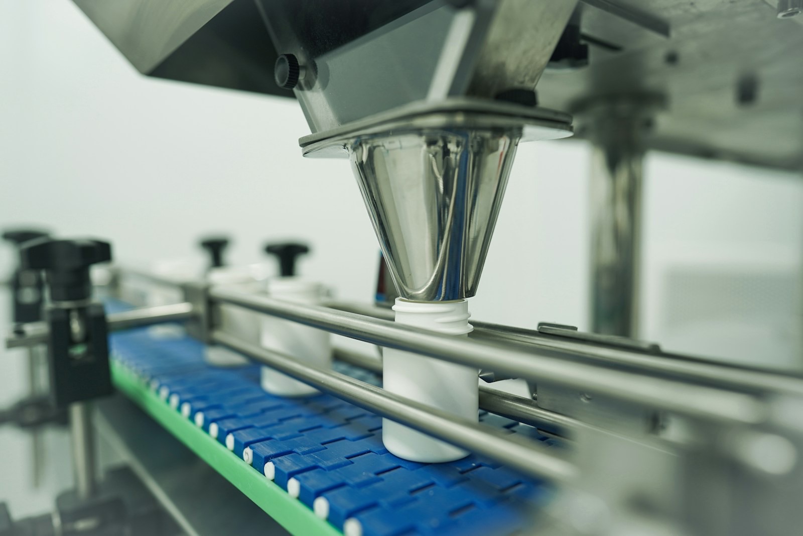 White plastic bottles moving along a blue conveyor under a stainless steel filling funnel on an automated packaging line