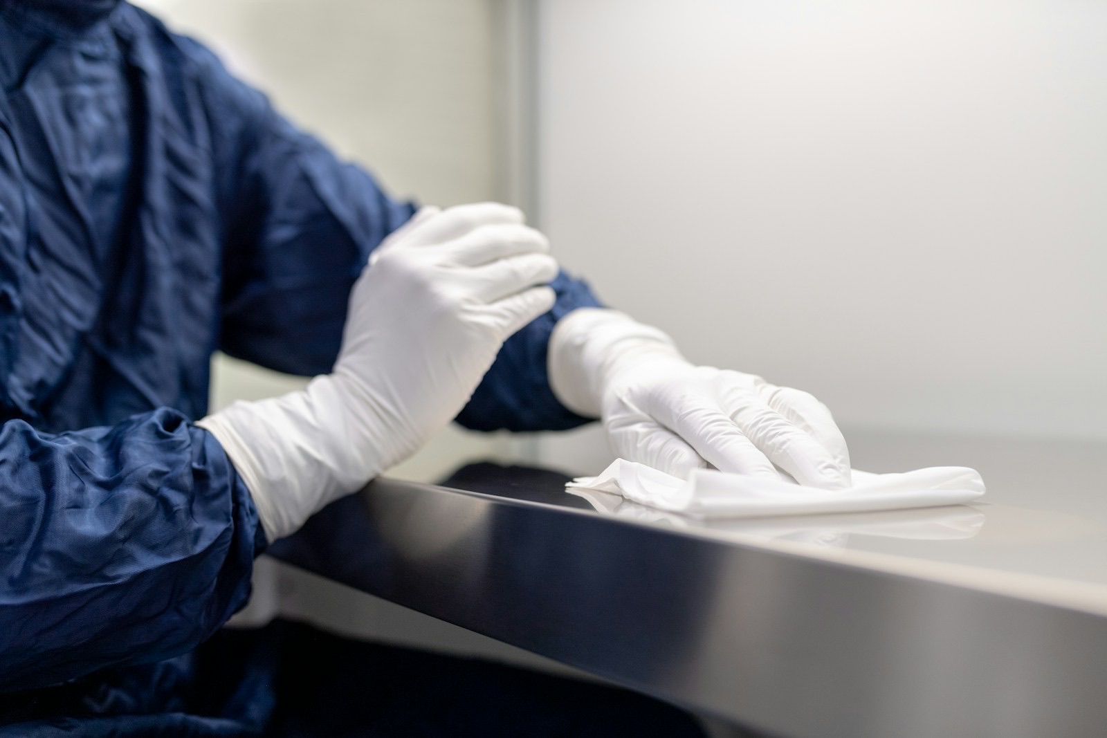 Gloved hands of a cleanroom operator in a blue gown wiping a stainless steel work surface