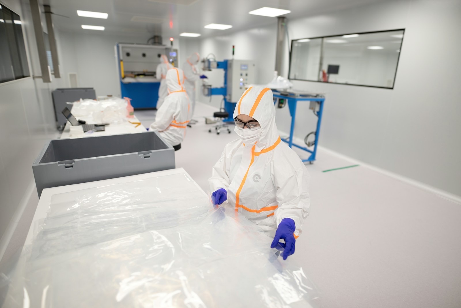 An operator in a white coverall, mask and goggles handling film at a bench in a cleanroom, with colleagues working at machines behind