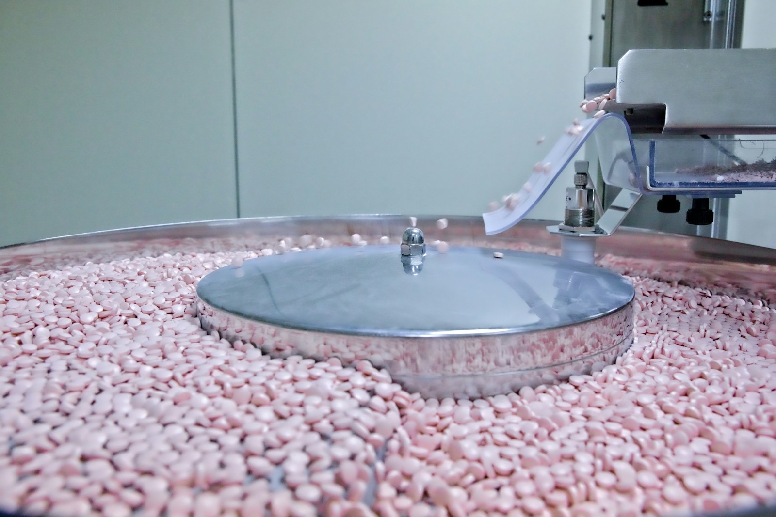 Pink tablets spilling from a stainless steel chute onto a rotating feeder bowl in a pharmaceutical production room
