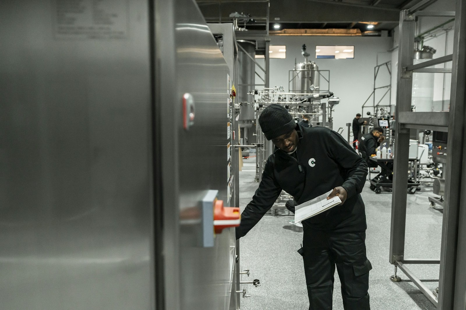 A technician holding a printed checklist reaches to inspect a stainless-steel equipment cabinet in a process manufacturing hall